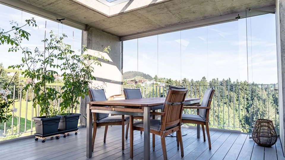 Loggia mit COVER Schiebe-Drehverglasung, Glasdach, Holzterrasse, Esstisch und Blick auf bewaldete Hügel