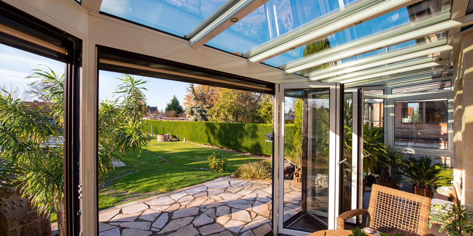 Wintergarten mit weisser FOLDER-Faltverglasung und Glasdach, Blick auf Rasen und Terrasse aus Natursteinplatten